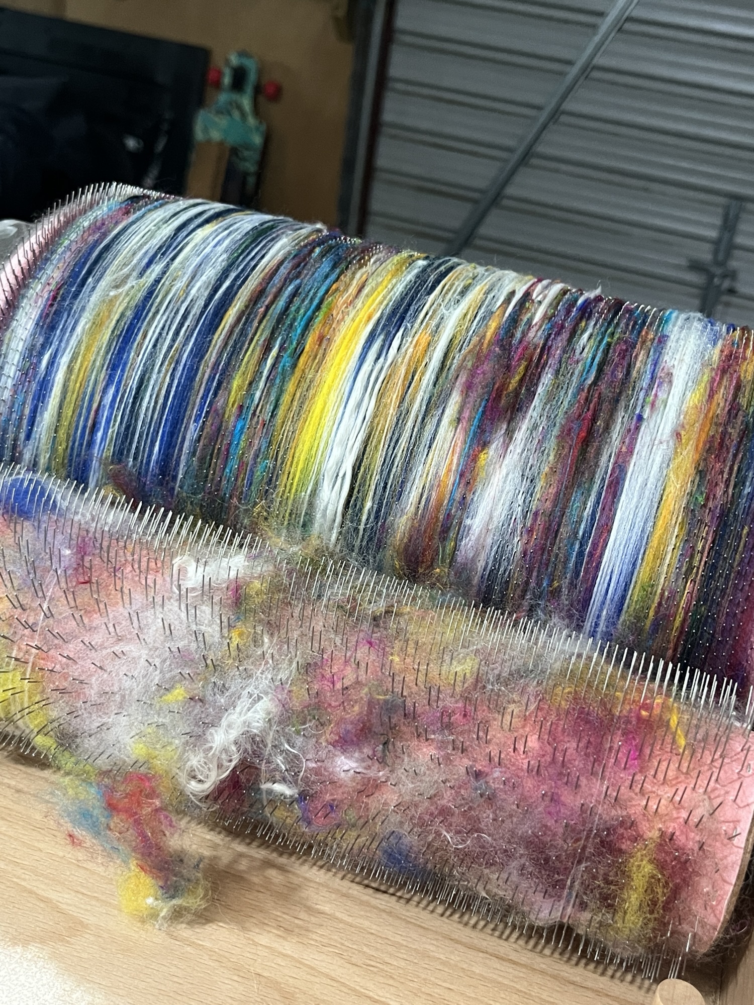 A blue batt for spinning or felting, curled up into a colourful dark blue spiral against a natural wood background. The batt is a fluffy blend of pure wool, silk and kid mohair with a dark blue base of NZ wool with pops of yellow wool and white kid angora mohair. This batt is ready to be handspun by handspinners on a spinning wheel or made into wool felt by felting