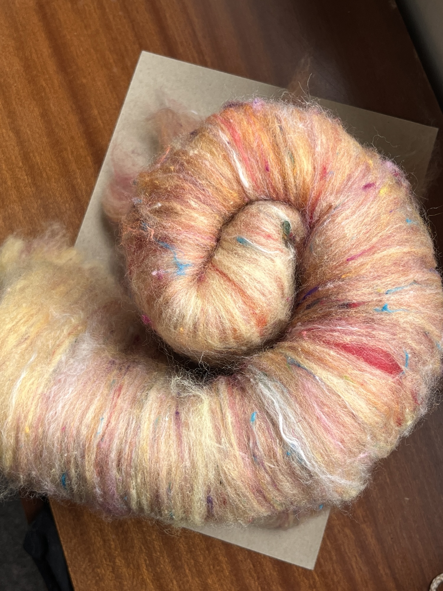 Handmade wool batt for spinning or felting: bright yellow NZ wool blended with sari silk, mulberry silk and kid mohair, featuring yellow and white accents, curled into a spiral on a natural wood background.