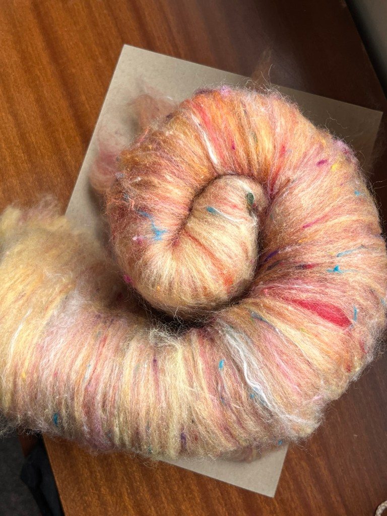 Handmade wool batt for spinning or felting: bright yellow NZ wool blended with sari silk, mulberry silk and kid mohair, featuring yellow and white accents, curled into a spiral on a natural wood background.