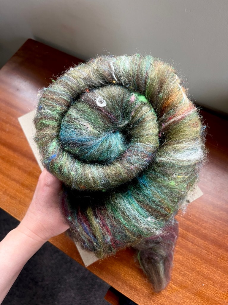 Handmade wool batt for spinning or felting: rich green and brown NZ wool blended with sari silk, mulberry silk and kid mohair, featuring yellow and white accents, curled into a spiral on a natural wood background.