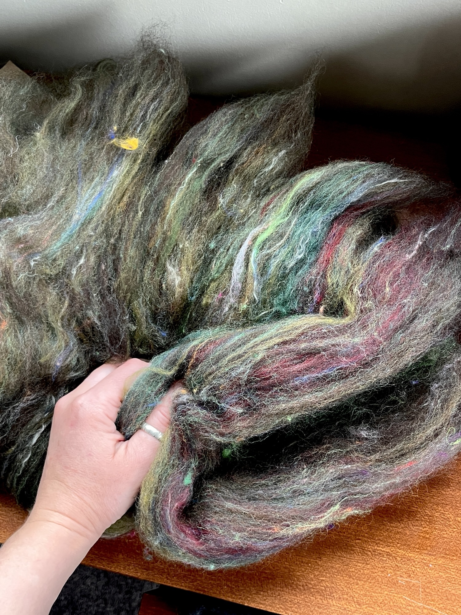 Handmade wool batt for spinning or felting: rich green and brown NZ wool blended with sari silk, mulberry silk and kid mohair, featuring yellow and white accents, curled into a spiral on a natural wood background.