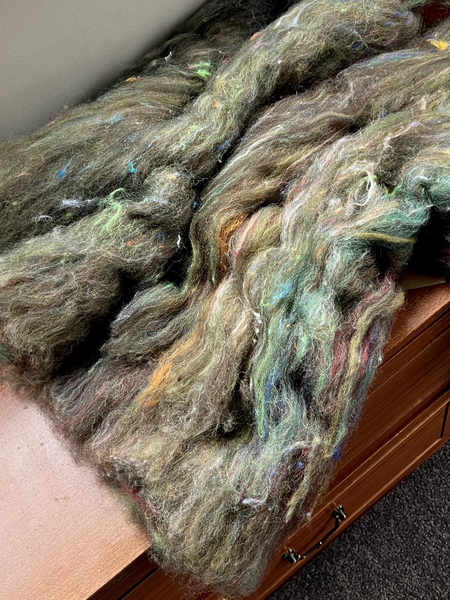 Handmade wool batt for spinning or felting: rich green and brown NZ wool blended with sari silk, mulberry silk and kid mohair, featuring yellow and white accents, curled into a spiral on a natural wood background.