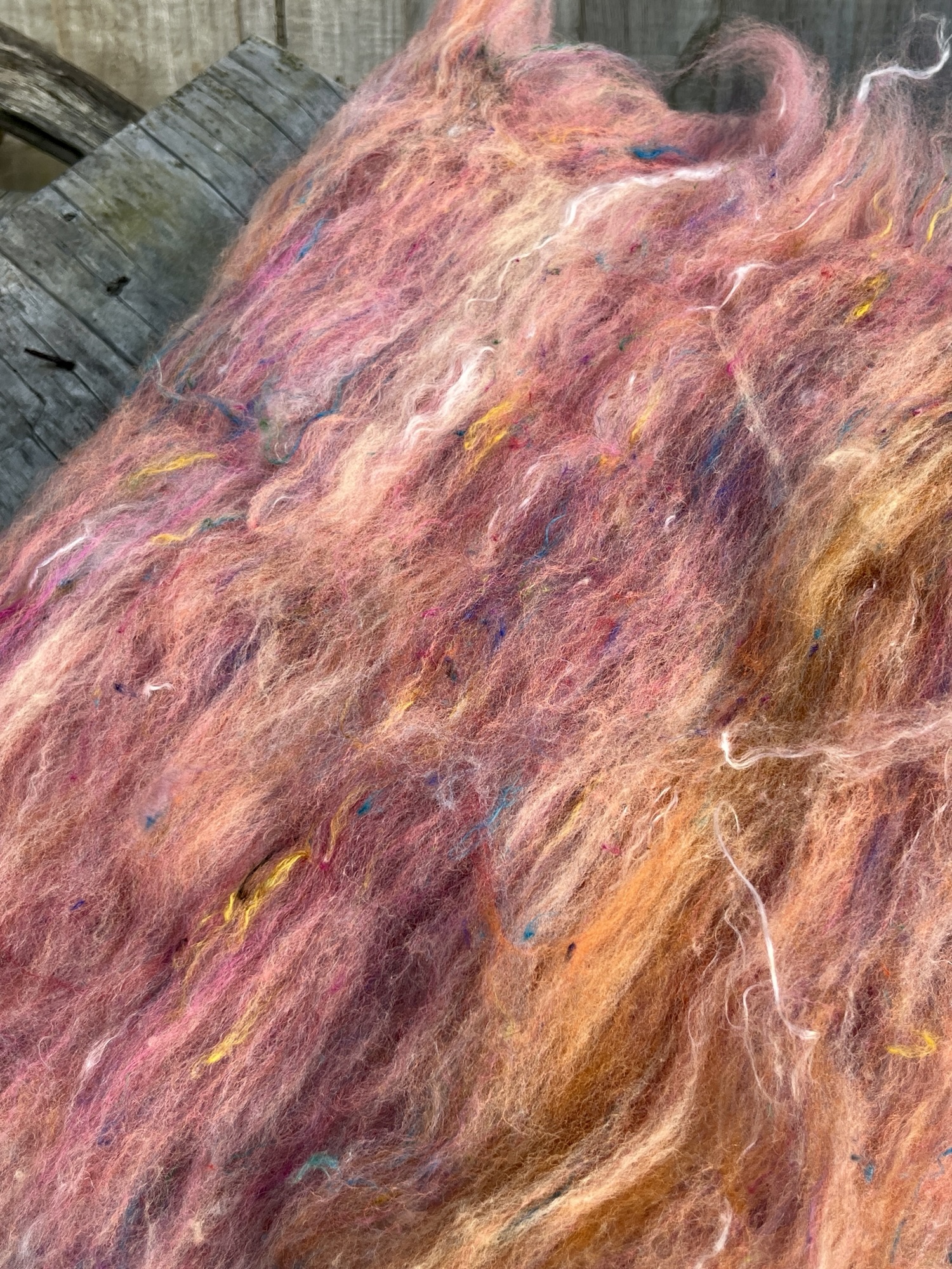 Handmade wool batt for spinning or felting: bright orange and yellow NZ wool blended with sari silk, mulberry silk and kid mohair, featuring yellow and white accents, curled into a spiral on a natural wood background.