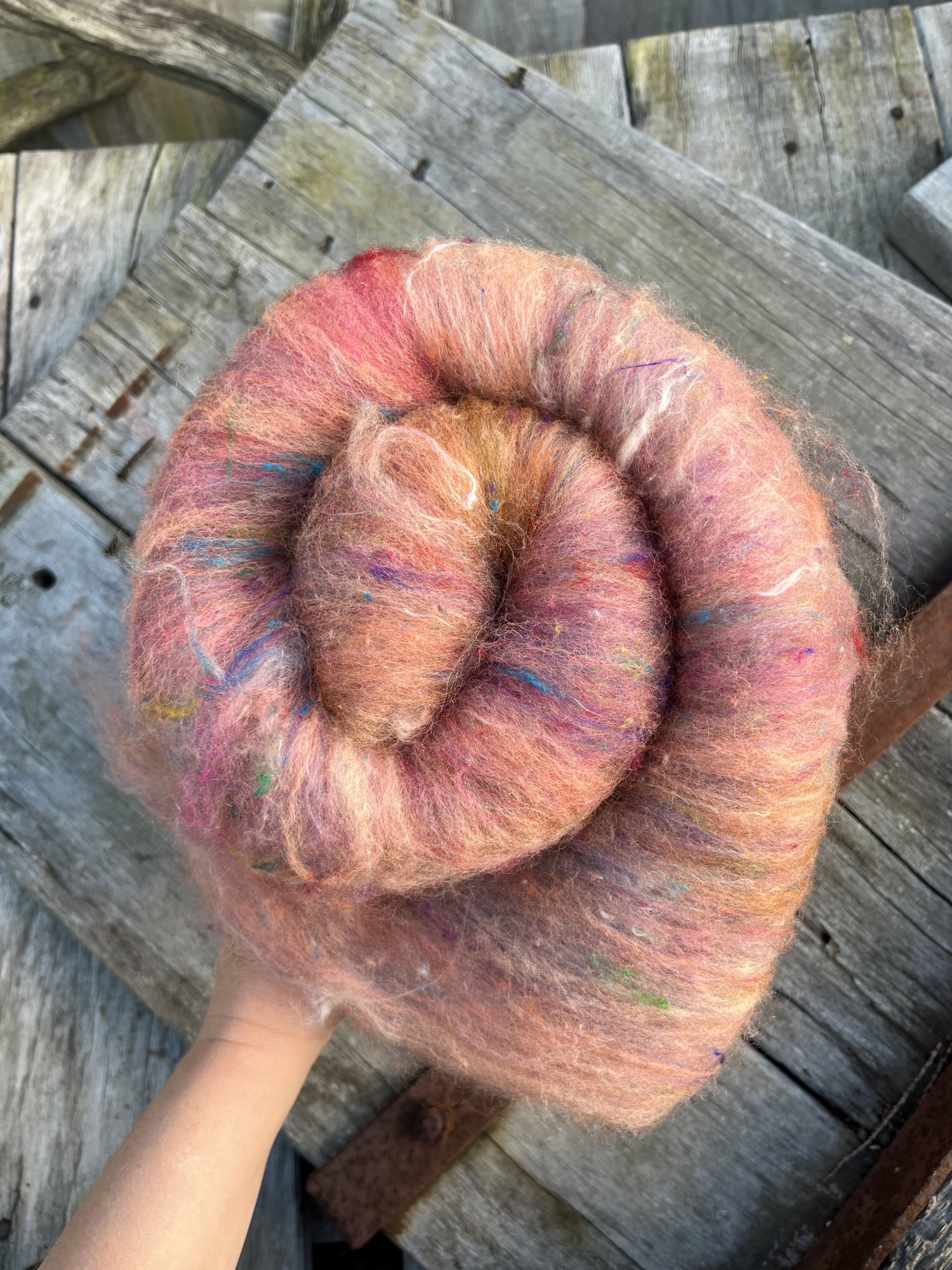 Handmade wool batt for spinning or felting: bright orange and yellow NZ wool blended with sari silk, mulberry silk and kid mohair, featuring yellow and white accents, curled into a spiral on a natural wood background.