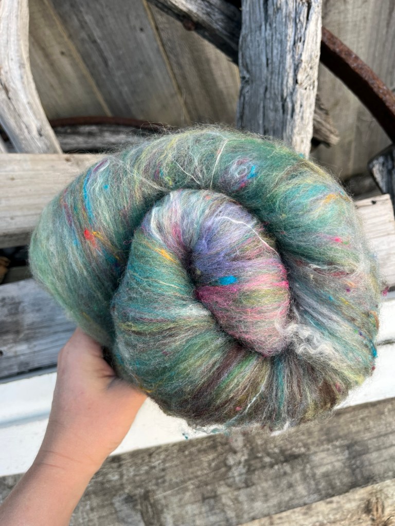 Handmade wool batt for spinning or felting: rich green NZ wool blended with sari silk, mulberry silk and kid mohair, featuring yellow and white accents, curled into a spiral on a natural wood background.