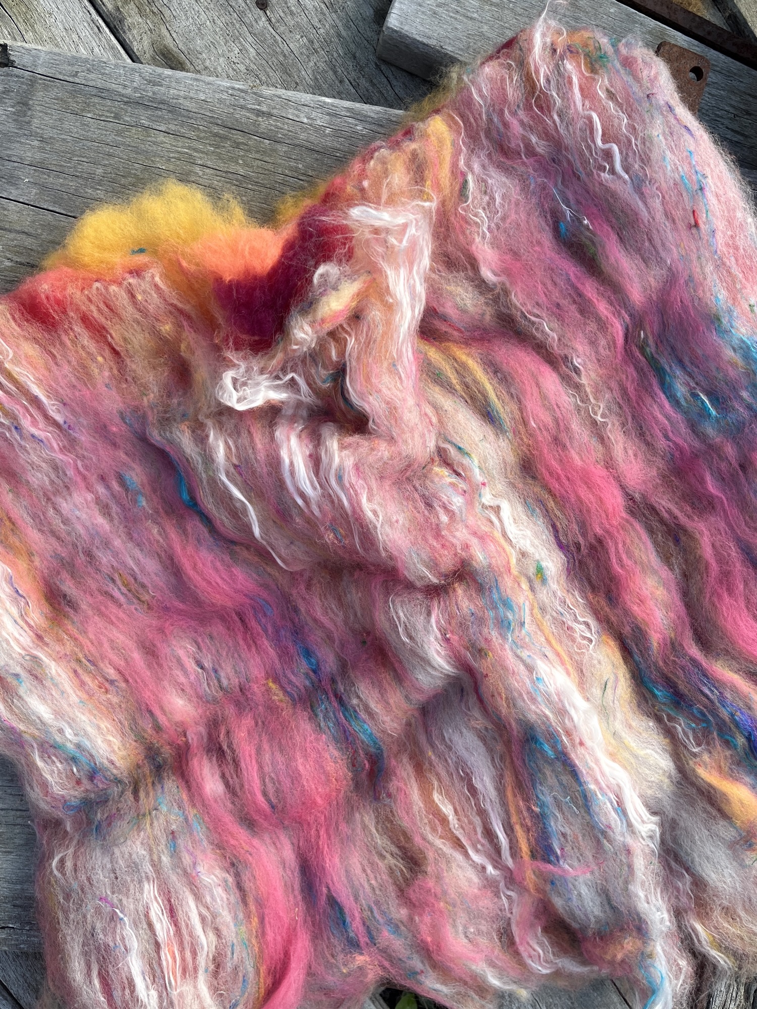 Handmade wool batt for spinning or felting: coral pink NZ wool blended with sari silk, mulberry silk and kid mohair, featuring blue and white accents, curled into a spiral on a natural wood background.