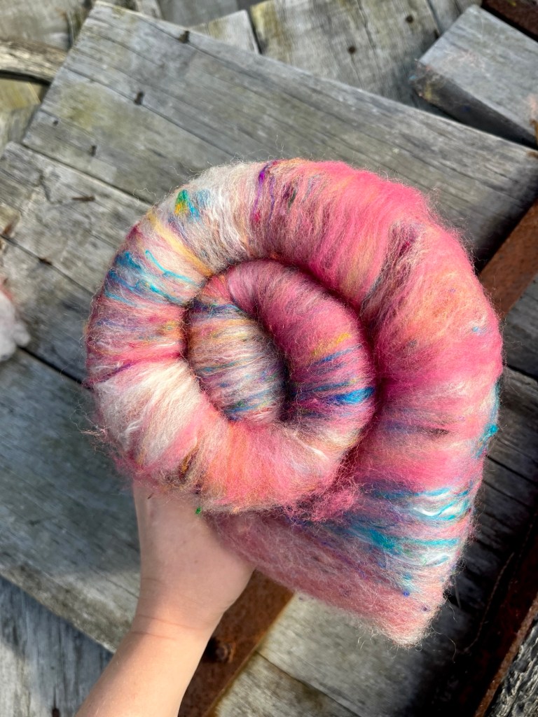Handmade wool batt for spinning or felting: coral pink NZ wool blended with sari silk, mulberry silk and kid mohair, featuring blue and white accents, curled into a spiral on a natural wood background.