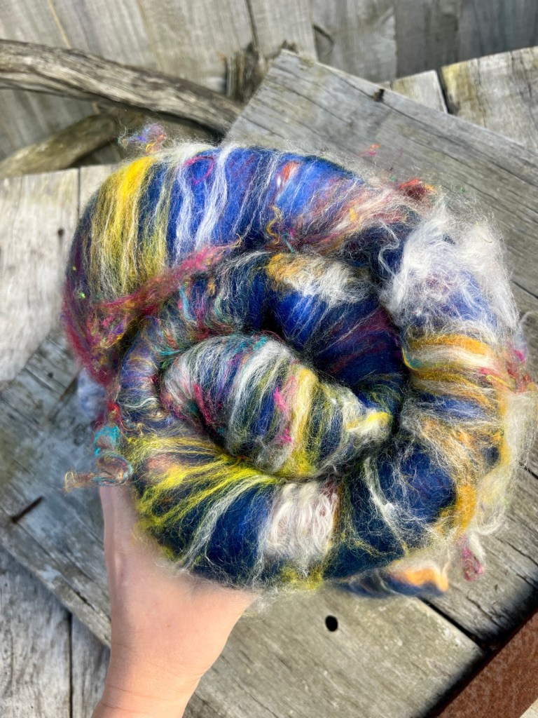 A blue batt for spinning or felting, curled up into a colourful dark blue spiral against a natural wood background. The batt is a fluffy blend of pure wool, silk and kid mohair with a dark blue base of NZ wool with pops of yellow wool and white kid angora mohair. This batt is ready to be handspun by handspinners on a spinning wheel or made into wool felt by felting