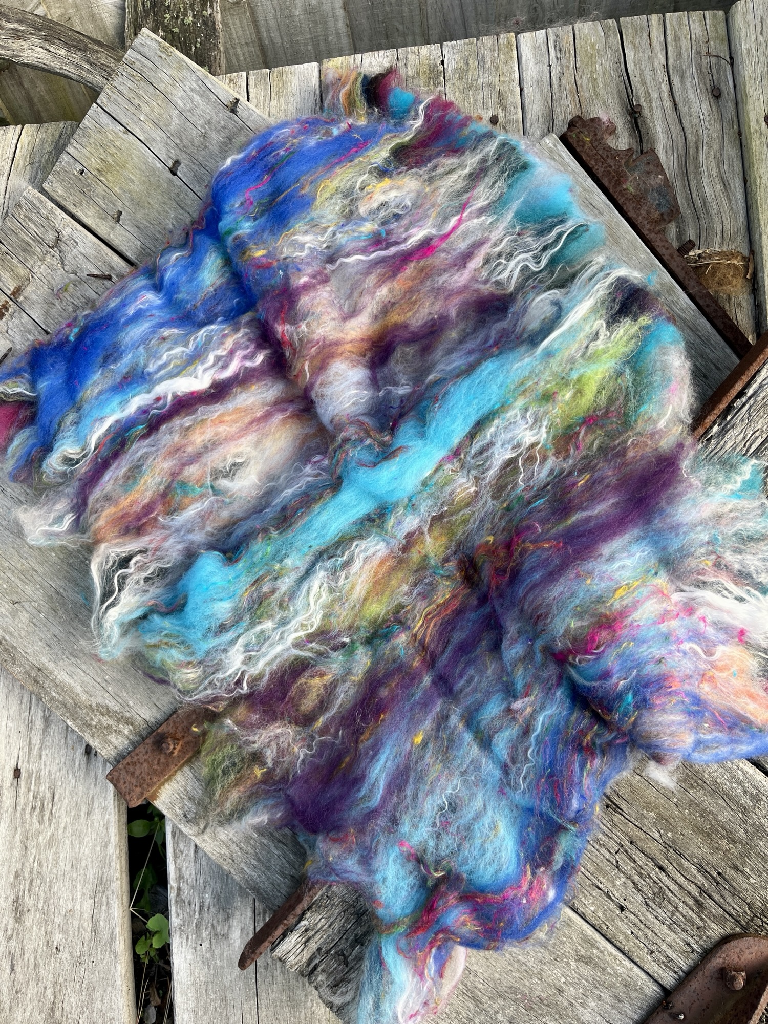 Handmade wool batt for spinning or felting: Turquoise blue NZ wool blended with sari silk, mulberry silk and kid mohair, featuring yellow and white accents, curled into a spiral on a natural wood background.