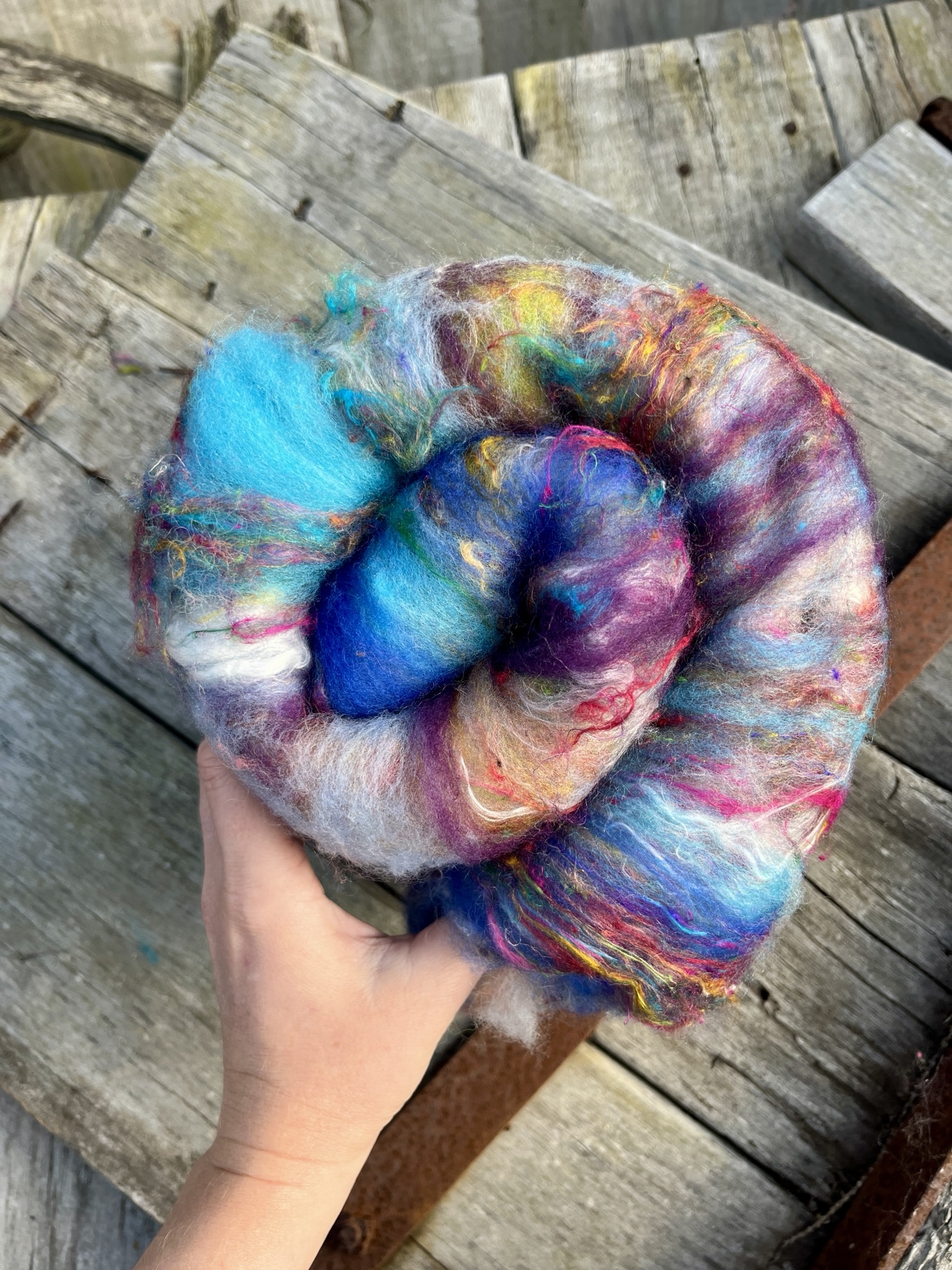 Handmade wool batt for spinning or felting: Turquoise blue NZ wool blended with sari silk, mulberry silk and kid mohair, featuring yellow and white accents, curled into a spiral on a natural wood background.