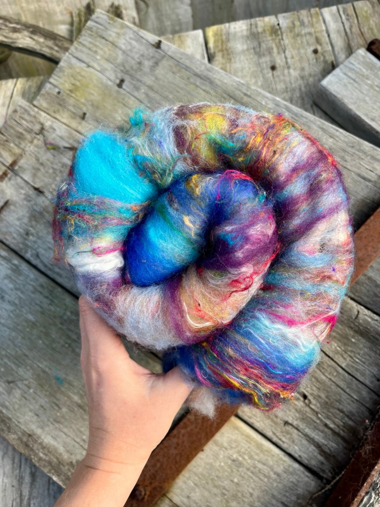Handmade wool batt for spinning or felting: Turquoise blue NZ wool blended with sari silk, mulberry silk and kid mohair, featuring yellow and white accents, curled into a spiral on a natural wood background.