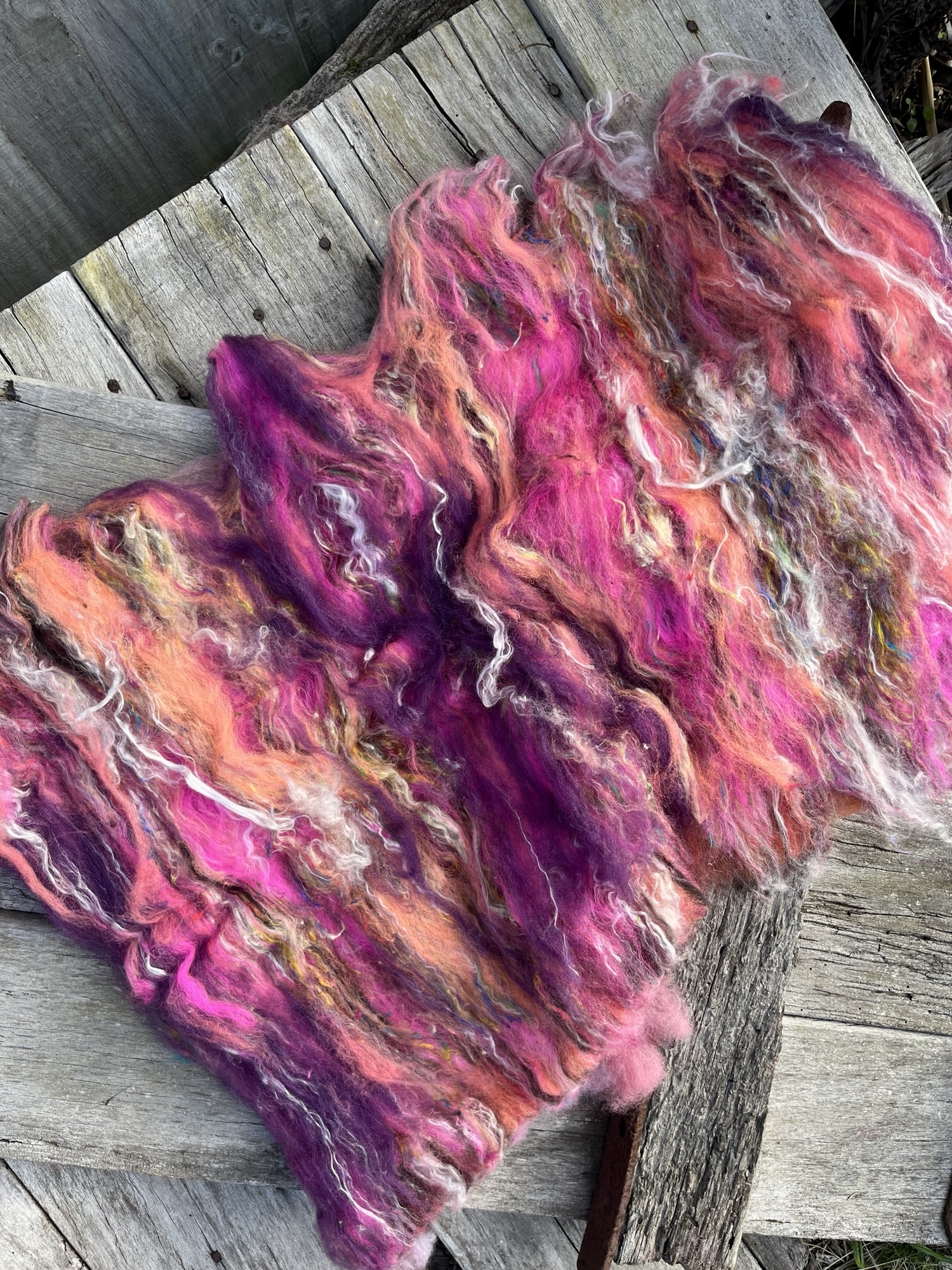 Handspun wool batt for spinning or felting: bright pink NZ wool blended with silk and kid mohair, featuring yellow and white accents, laid out on a natural wood background.