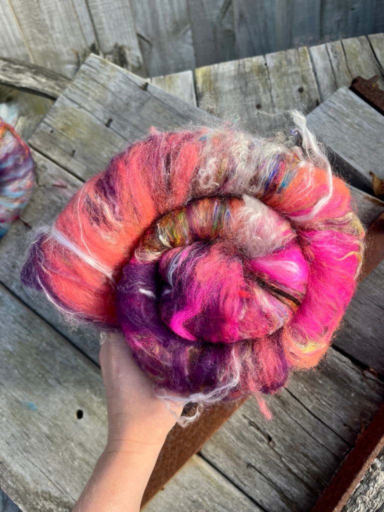 Handspun wool batt for spinning or felting: bright pink NZ wool blended with silk and kid mohair, featuring yellow and white accents, curled into a spiral on a natural wood background.