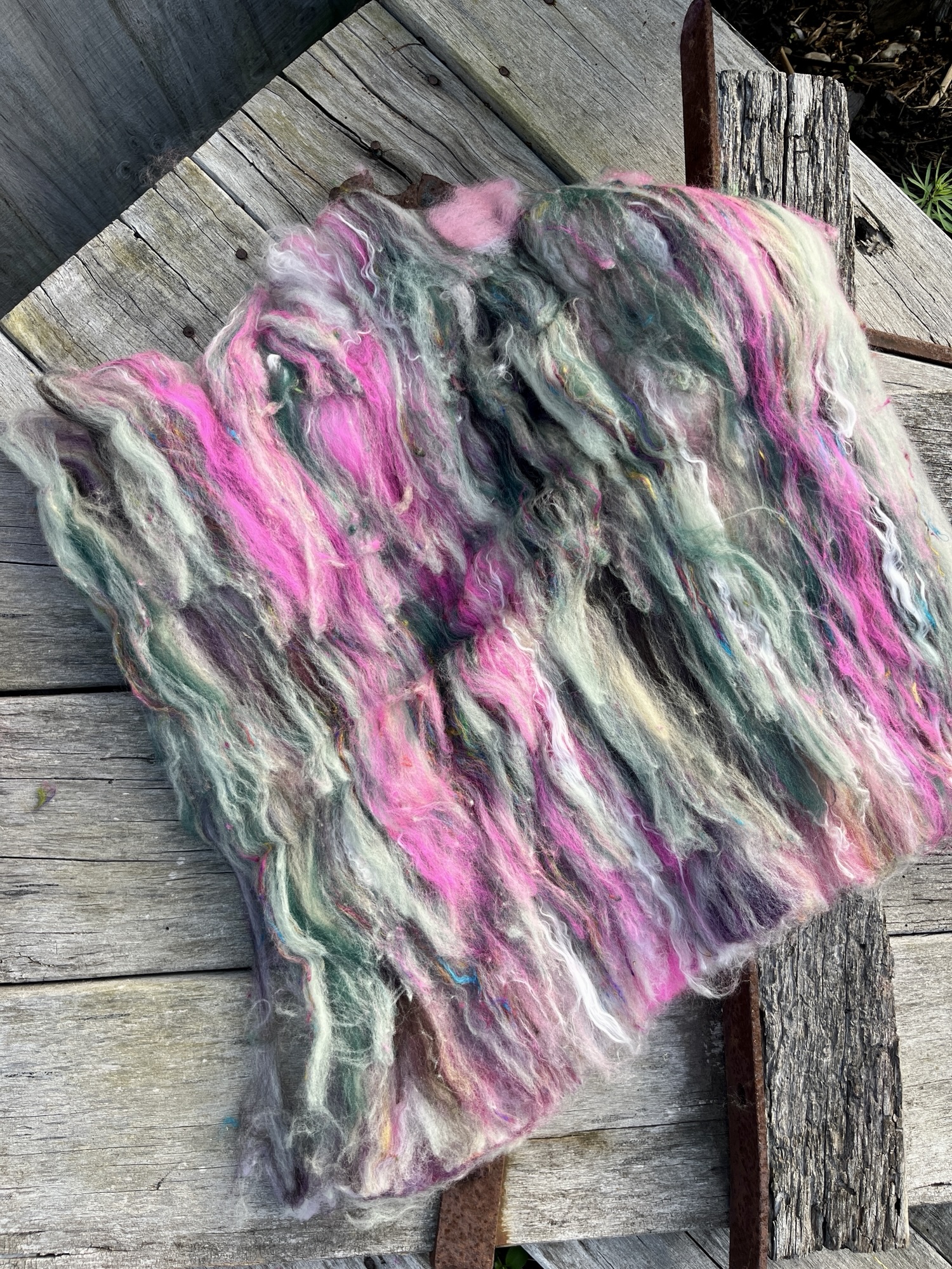 Handmade wool batt for spinning or felting: green and pink NZ wool blended with sari silk, mulberry silk and kid mohair, featuring yellow and white accents, curled into a spiral on a natural wood background.