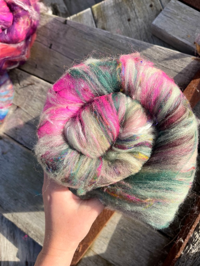 Handmade wool batt for spinning or felting: green and pink NZ wool blended with sari silk, mulberry silk and kid mohair, featuring yellow and white accents, curled into a spiral on a natural wood background.