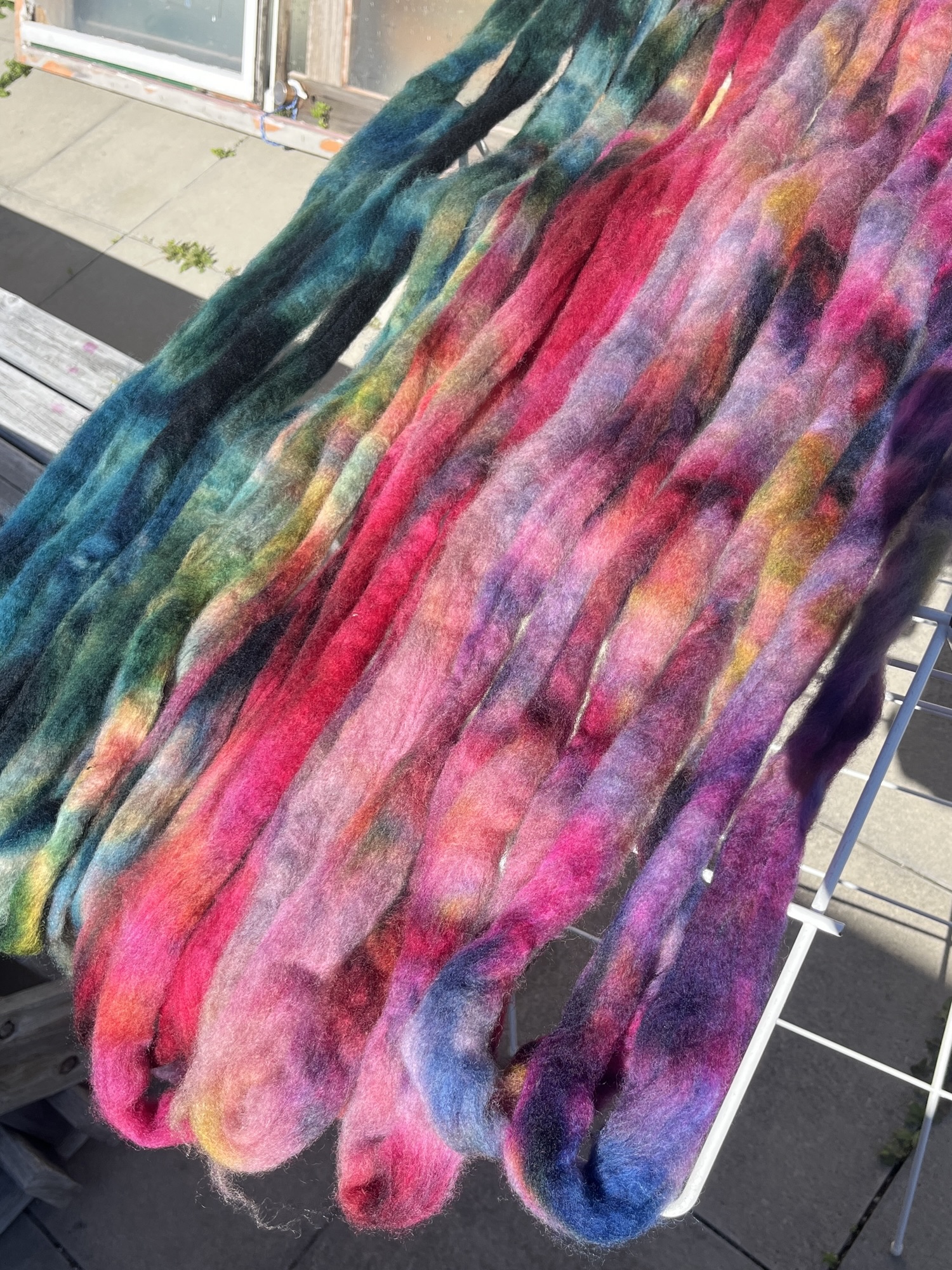 Strips of bright colored wool are laid across a clothesrack, drying in the sun. From left to right, a gradient moves through deep dark teal green trhough to bright pink and getting lighter into purple and eventually a dark purple with splotches of colour all over